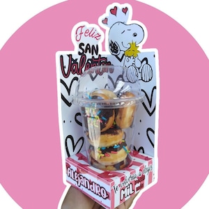 Può includere: Un biglietto rosa e bianco con il testo "Feliz San Valentin" e un'immagine di cartone animato di Snoopy che tiene un uccello. Davanti al biglietto c'è un bicchiere di plastica trasparente pieno di ciambelle ricoperte di cioccolato. Il bicchiere è appoggiato su una scatola rosa e bianca con il testo "Alejandro" e "Te volveria a elegir mil veces mas".