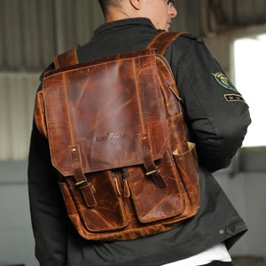 May include: A brown leather backpack with a flap closure and two front pockets. The backpack has a brand logo that says "SAINT STAG".