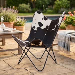 May include: A black and white cowhide butterfly chair with a black metal frame. The chair is positioned on a patio, with other furniture and plants in the background. The chair's seat has a distinctive spotted pattern.