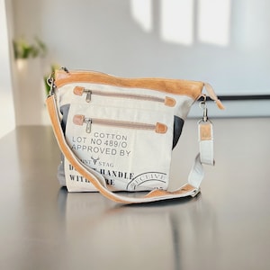 May include: A beige canvas tote bag with brown leather accents and a black strap. The bag has a zipper closure and a printed label that reads "Cotton Lot No 489/0 Approved By" with a stag logo. The strap has a metal clasp and a tag that reads "Receive".