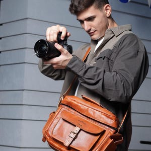 May include: A man wearing a brown and gray shirt is holding a black camera with a brown leather camera bag slung over his shoulder. The bag has a buckle and a brand name on the front.