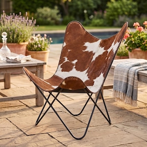 May include: A brown and white cowhide butterfly chair with a black metal frame. The chair is positioned on a stone patio, with plants and a pool visible in the background. The chair's design resembles a butterfly's wings.