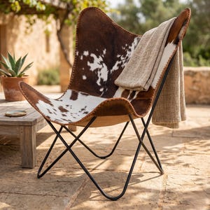 Könnte beinhalten: Ein braun-weißer Butterfly-Stuhl aus Kuhfell mit einem schwarzen Metallrahmen. Eine beige Decke ist über die Rückenlehne des Stuhls drapiert. Der Stuhl steht im Freien in der Nähe eines Holztisches und einer Topfpflanze.