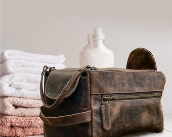 May include: A brown leather travel toiletry bag with a zipper closure and a strap. The bag is sitting on a white surface next to a stack of towels and a clear bottle.