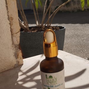 Peut inclure: Une bouteille en verre brun avec un bouchon doré et un compte-gouttes. La bouteille est étiquetée "Cactus Oil" en arabe et en anglais. La bouteille est posée sur une surface gris clair.