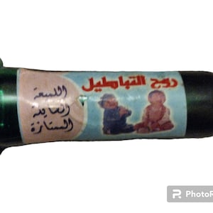 May include: A small green glass bottle with a black cap and a white label. The label has Arabic text and a picture of two children. The text on the label reads "Dukh Al-Tawafil" and "Wal-Salam Wa-Rahma".