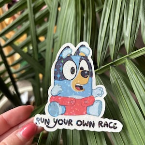 May include: A sticker featuring a cartoon dog character in blue, tan, and red. The dog has large, sparkling eyes and is sitting with its arms outstretched. The sticker has the text "RUN YOUR OWN RACE" in a handwritten style font.