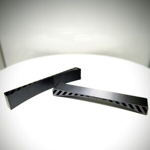 May include: Two black, rectangular objects with angled edges and diagonal cutouts. One is curved, the other straight. The objects are on a white surface. The cutouts are in a striped pattern.