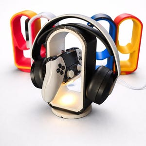 Può includere: Un supporto per cuffie e controller di gioco in bianco e nero con luce integrata. Il supporto contiene un controller bianco e nero. Il supporto è circondato da supporti ad arco colorati in rosso, giallo, bianco e blu.
