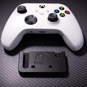 Può includere: Un controller Xbox bianco con joystick e pulsanti neri e un supporto per controller nero. Il supporto presenta il testo "Rinitynator" e un logo a forma di testa aliena. Il controller ha pulsanti colorati.
