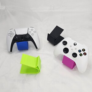 Könnte beinhalten: Ein weiß-schwarzer PlayStation 5-Controller und ein weißer Xbox-Controller, jeweils auf einem farbigen Ständer. Die Ständer sind blau, schwarz, grün und pink. Die Controller befinden sich auf weißem Hintergrund.