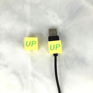Może przedstawiać: Dwie żółte osłony na pendrive z napisem "UP" w zielonych literach. Jedna osłona leży na białym tle, a druga jest przymocowana do czarnego kabla USB.