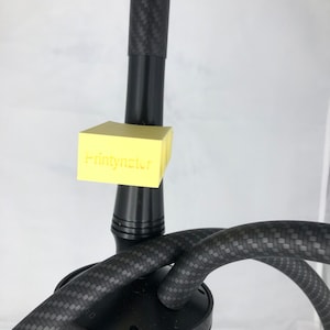 Könnte beinhalten: Schwarze Wasserpfeife mit Karbonfaser-Design. Die Wasserpfeife hat eine schwarze Basis und einen schwarzen Schlauch mit Karbonfaser-Muster. Ein gelber Aufkleber mit dem Text "Printynstr" ist an der Wasserpfeife befestigt.