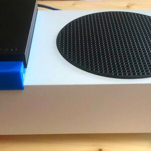 Könnte beinhalten: Eine weiße Spielekonsole mit schwarzer Oberseite und kreisförmigem Lüfter. Ein schwarzes Gerät liegt obenauf, unterstützt von blau, rot und grün gefärbten Erhöhungen. Das Xbox-Logo ist sichtbar.