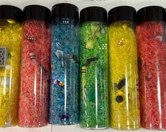 May include: Eight clear plastic bottles filled with different colored rice and small objects. The bottles are arranged in a row from left to right. The colors of the rice are green, yellow, red, blue, green, yellow, red, and blue.