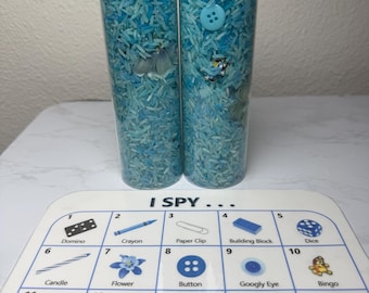 May include: Two clear plastic bottles filled with blue rice and various small objects. A game card with the text "I SPY..." and a grid of 15 squares with images and names of objects. The objects include a domino, crayon, paper clip, building block, dice, candle, flower, button, googly eye, bingo, red balloon, flower, bluey, rainbow cloud, and watermelon.
