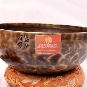 May include: A brown singing bowl with a hammered finish and a symbol on the side. The bowl is sitting on a cushion with a floral pattern. The label on the bowl reads "Svadhisthana Sacral Chakra D".