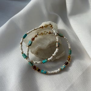 Puede incluir: Dos pulseras de cuentas con cuentas blancas y detalles turquesa y marrón. Las pulseras están superpuestas.