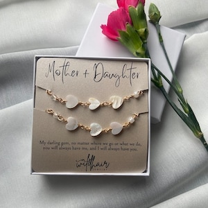 May include: A gift set featuring two gold-toned bracelets with heart-shaped charms, presented in a box with the text "Mother + Daughter." A pink carnation flower rests beside the box on a gray surface.