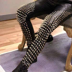 Puede incluir: Pantalones de cuero negro con tachuelas plateadas que cubren las piernas. Los pantalones los lleva puestos una persona sentada en una silla.