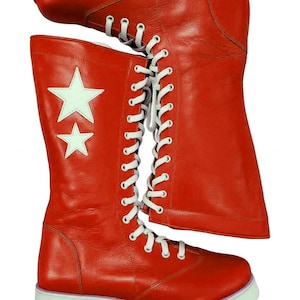 Puede incluir: Botas de cuero rojas con detalles de estrellas blancas y cordones blancos. Las botas tienen una suela blanca con una franja morada. Las botas son hasta la rodilla y tienen un diseño clásico con cordones.