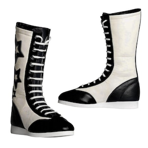 Pode incluir: Par de botas altas de duas cores. As botas são principalmente brancas com detalhes em preto na biqueira, calcanhar e acabamento superior. Uma bota apresenta recortes em forma de estrela. Cadarços brancos sobem pela frente de cada bota.