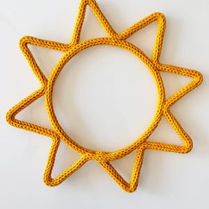 Könnte beinhalten: Gelb-goldene Wanddekoration in Sonnenform. Das Sonnendesign besteht aus dickem, strukturiertem Garn mit einem kreisförmigen Zentrum und nach außen gerichteten Strahlen. Das Stück befindet sich vor einem weißen Hintergrund.