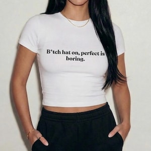 Op de afbeelding: Witte crop top met de tekst "B*tch hat on, perfect is boring." Het shirt heeft korte mouwen en wordt gecombineerd met een zwarte joggingbroek. Een gouden ketting en armbanden zijn ook zichtbaar.