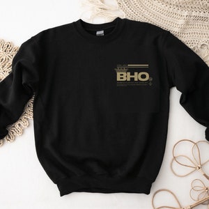 Op de afbeelding: Zwart sweatshirt met een gouden kroon en de tekst "BHO" bedrukt op de voorkant.