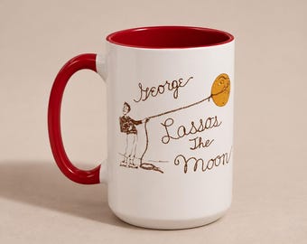 May include: A white ceramic mug with a bright red interior and handle. The exterior features a brown line drawing of a person lassoing a yellow moon, accompanied by the brown cursive text that reads George Lassos The Moon in a clear font.
