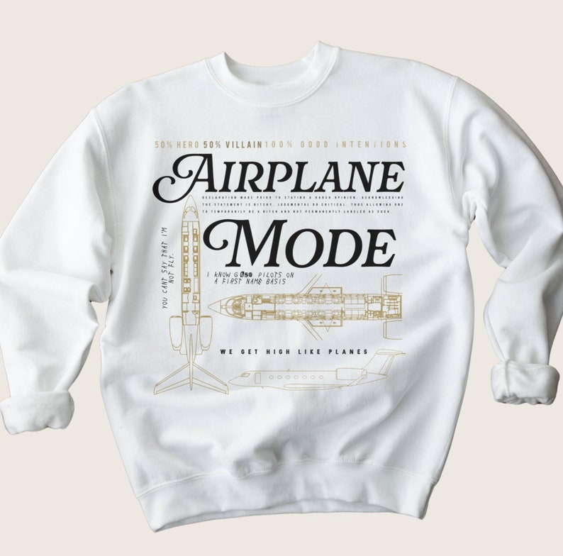 Op de afbeelding: Witte trui met een gouden vliegtuiggrafiek en de tekst "50% Hero 50% Villain 100% Good Intentions Airplane Mode" en "We get high like planes".
