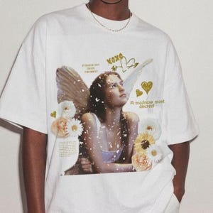 May include: White t-shirt featuring a graphic of a woman with angel wings, flowers, and text. The design includes the words "XOXO" and "A madness most discreet" in gold. The shirt is a casual, unisex style.