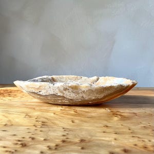 May include: A natural stone bowl with an irregular shape. The bowl has a mix of beige, tan, and cream colours, with a textured exterior and a smooth interior. The bowl is displayed on a wooden surface.