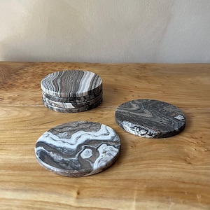 May include: A stack of six and two individual round coasters. The coasters have a marbled pattern in shades of gray, brown, and white. They are made of a stone-like material and sit on a wooden surface.