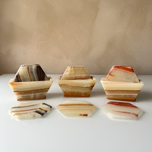May include: A set of onyx coasters and matching holders. The coasters and holders are hexagonal and feature natural bands of brown, tan, and white. The holders have a boat-like shape. The set includes three holders and three coasters.