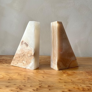 May include: Two triangular bookends. One is white with brown and tan marbling, the other is tan with light brown veining. The bookends are made of stone and sit on a wooden surface.