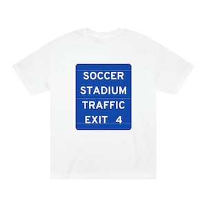 May include: White t-shirt with a blue square graphic. The graphic reads "SOCCER STADIUM TRAFFIC EXIT 4" in white text. The shirt is a casual style, suitable for everyday wear.
