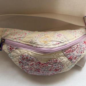 May include: A quilted fanny pack featuring a patchwork design of cream, pink, and yellow floral fabrics. The bag includes a light purple zipper closure, a wide cream-colored adjustable strap, and iridescent metal hardware attachments on each side.