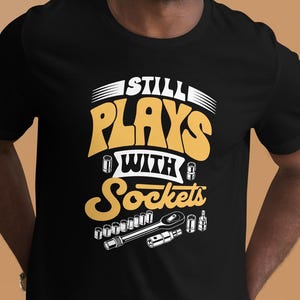 Könnte beinhalten: Schwarzes T-Shirt mit dem Text "Still Plays With Sockets" in Weiß und Gelb. Das Design beinhaltet Werkzeugillustrationen. Das T-Shirt wird von einer lächelnden Person getragen.