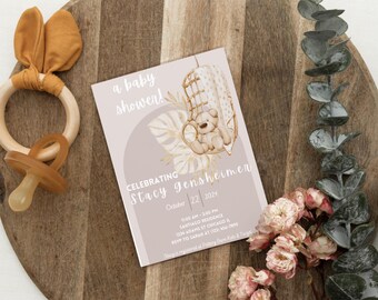 May include: A baby shower invitation with a watercolor illustration of a teddy bear sitting in a hanging chair. The invitation is on a light pink background with gold accents and the text "a baby shower! Celebrating Stacy gensheimer October 22, 2024"