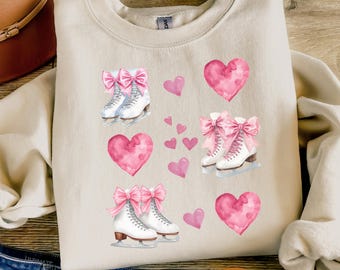 May include: A sand-colored sweatshirt featuring a design of pink hearts and white ice skates with pink bows. The word "Sand" is printed on the bottom left corner of the image. The sweatshirt is folded on top of blue jeans.
