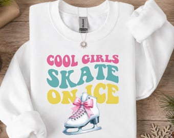 May include: A white long-sleeve crewneck sweatshirt featuring the text COOL GIRLS SKATE ON ICE in pink, teal, and yellow wavy lettering. Below the text is a graphic of a white ice skate with pink laces and a pink bow.