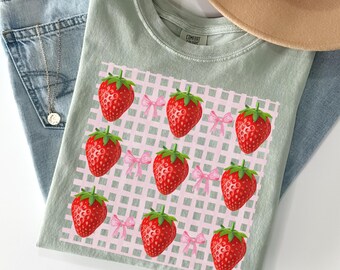 May include: A light green t-shirt featuring a strawberry design. The design includes red strawberries with green tops and pink bows on a pink and white checkered background. The shirt is folded on top of blue jeans.