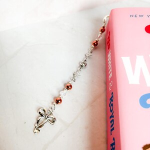 May include: A silver bookmark with a bow charm and a chain of alternating copper and clear beads. The bookmark is attached to a pink book with the text "NEW YORK WHITE & ROYAL BLUE" on the spine.