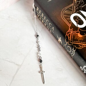 May include: A silver bookmark with a sword charm and black and clear beads. The bookmark is hanging from a book with the title "One Dark Window" by Rebecca Yarros.
