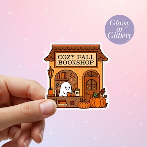 May include: A sticker featuring a cozy fall bookshop illustration. The shop has an orange facade, a brown roof, and a ghost reading a book. Pumpkins and fall leaves decorate the scene. The text "COZY FALL BOOKSHOP" is displayed.