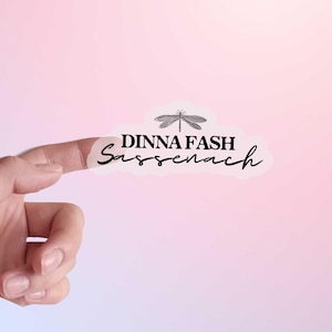 May include: A finger pointing to a white sticker with a dragonfly and the text "Dinna Fash Sassenach".