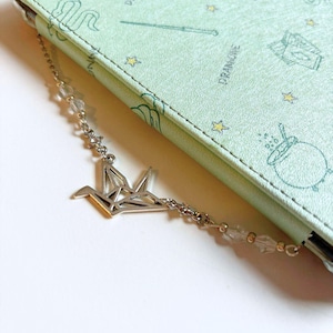 May include: A silver origami crane charm hangs from a silver chain with clear beads. The chain is attached to a green book with a whimsical pattern of stars, wands, and other magical objects.