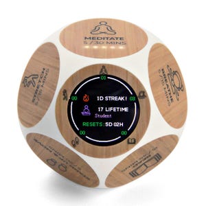 May include: A white and wood-grain cube-shaped device with a digital display. The display shows text and icons related to wellness activities. The cube has labels for activities like meditation, stretching, and breathing. The device tracks streaks and lifetime achievements.