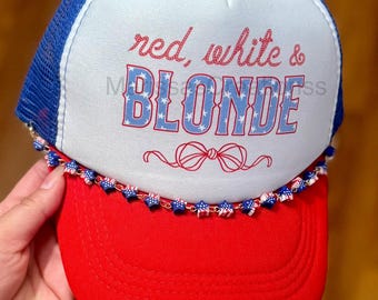 May include: A trucker hat with a white front panel, blue mesh back, and a red brim. The front features the red text "red, white &" above the blue text "BLONDE" with a star pattern, and a red line-art bow. A chain of small star-shaped beads lines the brim.